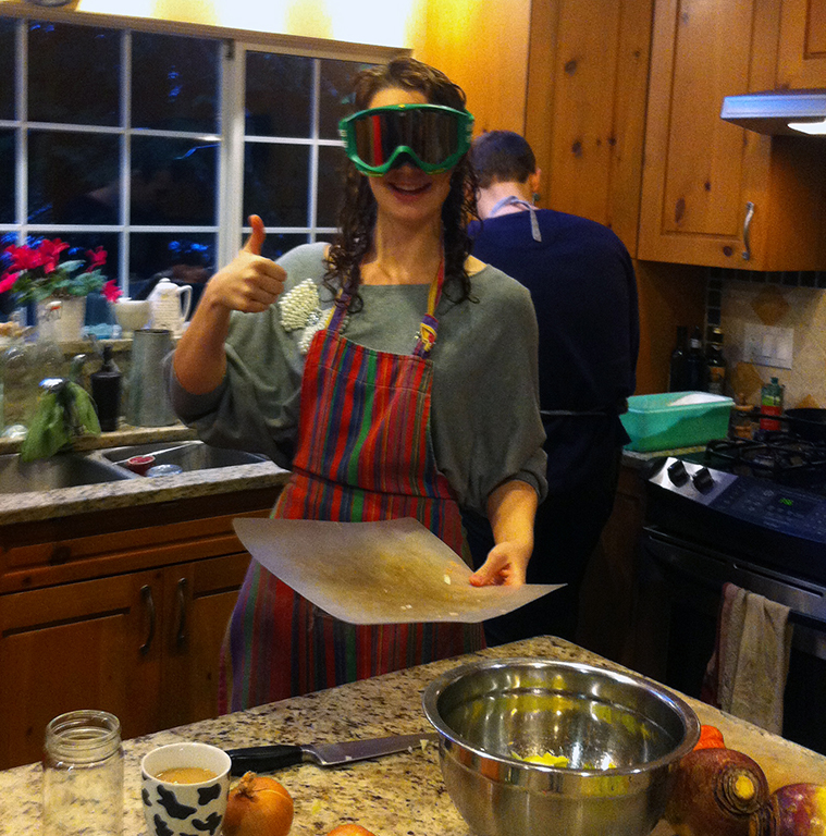 Michelle Gamage, wearing ski goggles and a colourful striped apron, gives the camera the thumbs up. She’s holding a plastic cutting board, and there’s a metal bowl and some onions on the counter in front of her.
