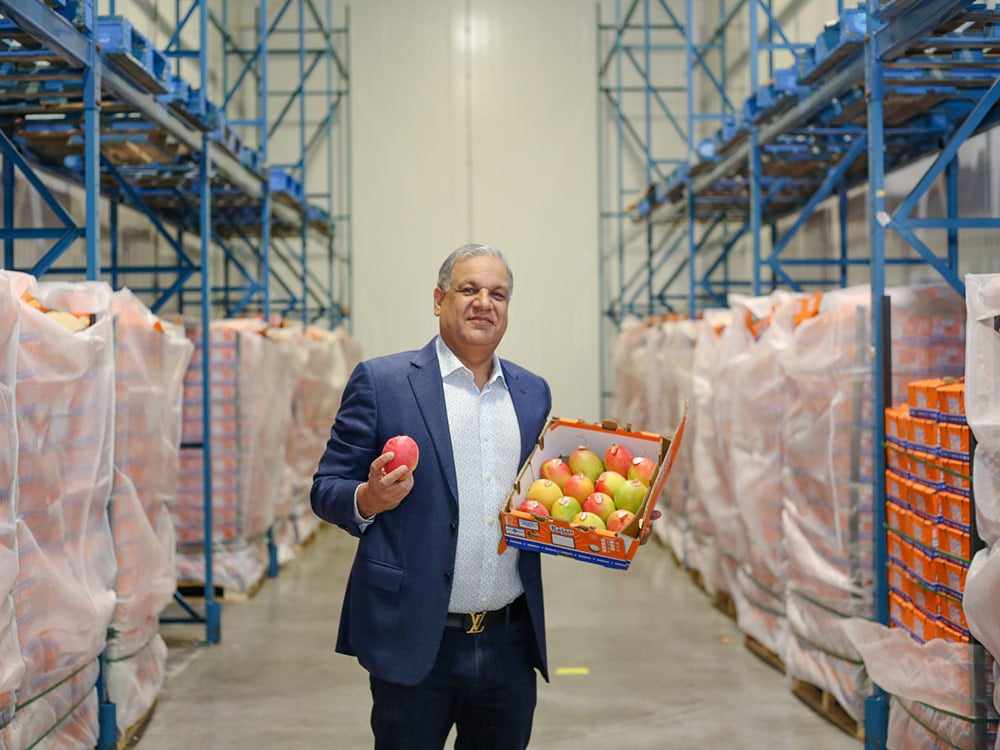 Tony Singh wears a blue blazer over a white dress shirt with floral texture. He is standing in the middle of a warehouse. On either side of him are blue shelves holding pallets of Singh’s signature Rajan brand Haden mangoes. The orange flats are ensconced in white coverings. In his left arm, Singh is holding an orange box of the mangoes. They have red, orange, yellow and green flesh. Singh holds up one mango for the camera with his right hand. He is smiling.