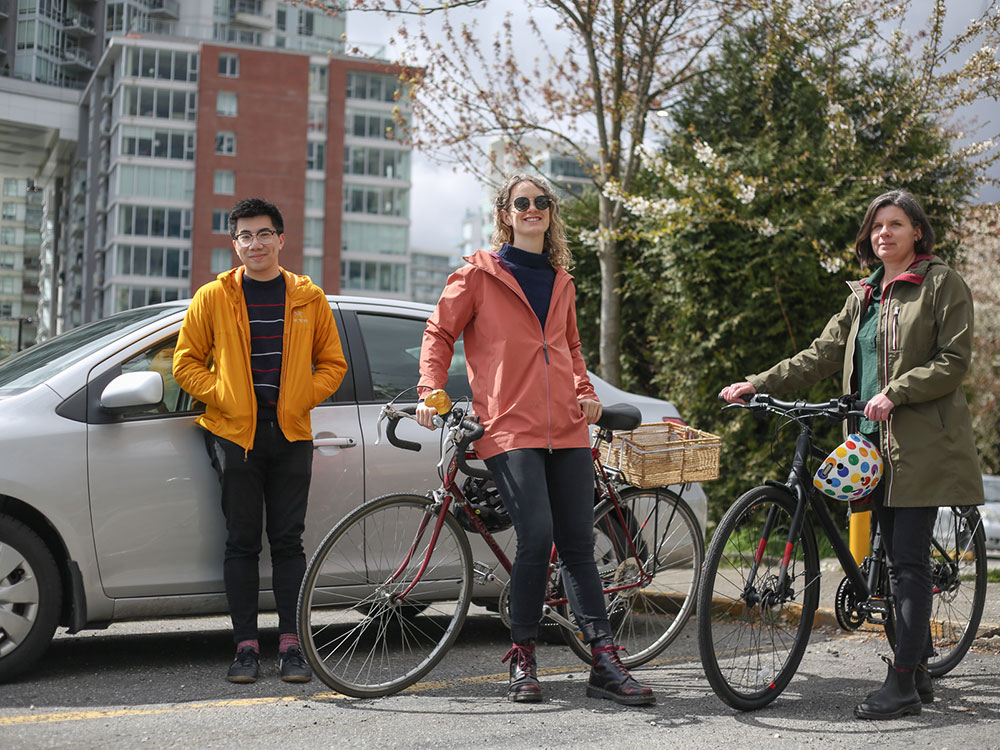 Three journalists ranging in age from 25 to 40 stand with their modes of transport, a car and two bikes.