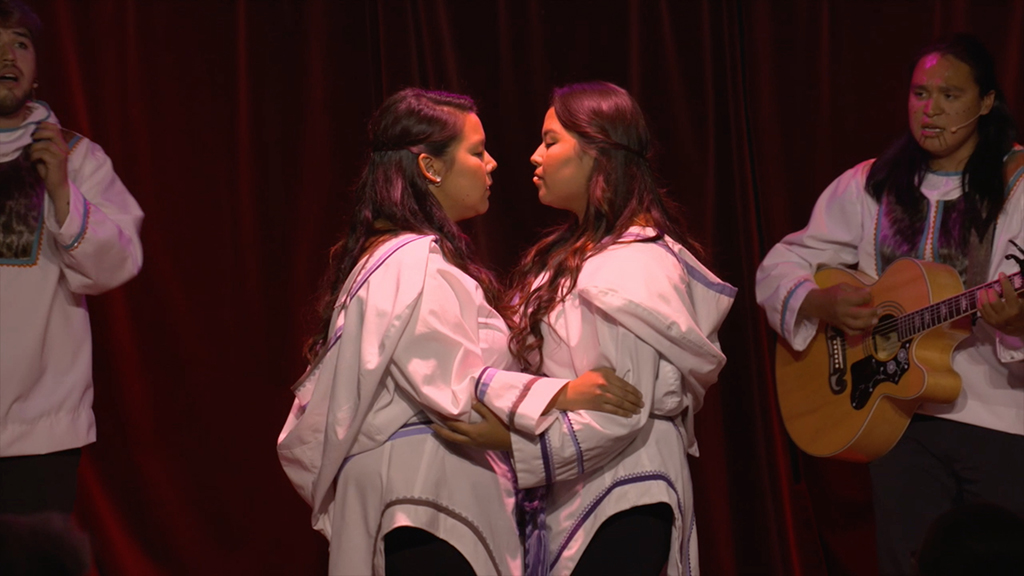 Leanna Wilson, left, and Tooma Laisa, right, stand facing each other, holding each other’s arms. They are wearing traditional white tops with blue trim. They are lit with stage lighting, and the background is black. Behind them to the left and right, two people wearing similar white shirts are singing. One is playing guitar.