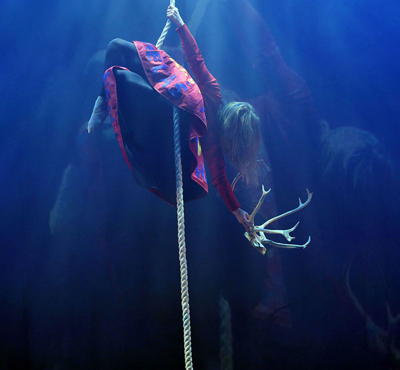 Camilla Therese Karlsen wears a red patterned dress, black tights and ballet shoes. Her long brown hair obscures her face. She hangs from a rope running down the middle of the photograph. She is holding an antler. Behind her is a dark blue background. The scene is lit with stage lighting.