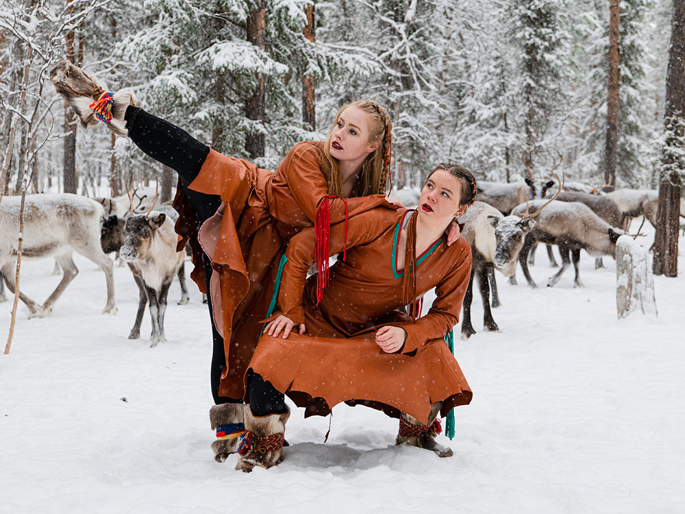Two women are wearing matching dresses the colour of terra cotta. Liv Aira kicks her right foot into the air as she rests her arms across Marika Renhuvud’s shoulders. They are both looking to the left of the camera, their expressions serious. They are standing in white snow. Behind them is a snowy forest and flock of reindeer.