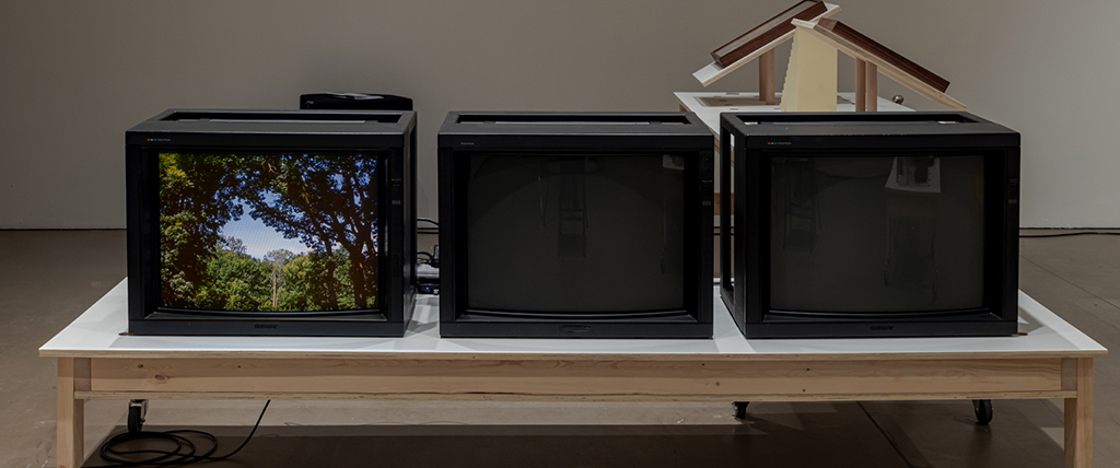 Three black television screens sit next to each other on a low birch table. The screen on the left features a sunny image of green trees against a blue sky. The other screens are blank and turned off. In the background, two unseen pieces of artwork are on display on table stands.