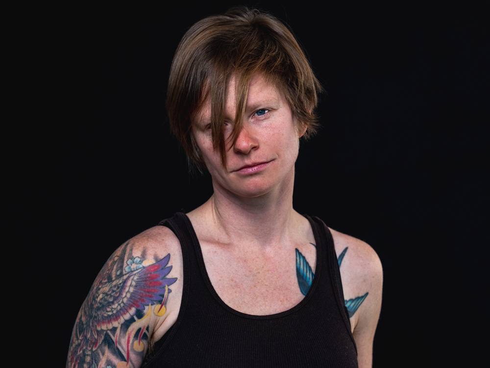 An author photo of Lori Fox. Lori is a white non-binary person wearing a black tank top. Colourful tattoos are visible on their arms and shoulders. 