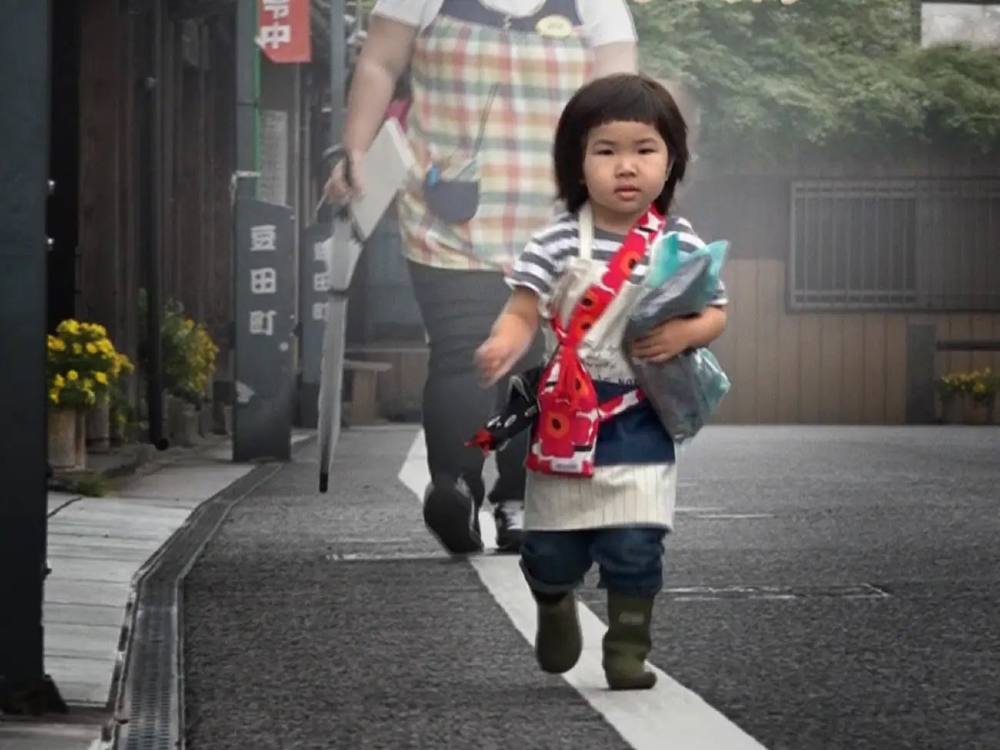 A toddler is walking down a street in Japan. She has short black hair with baby bangs and is wearing an apron over a striped T-shirt. She has a purse in red floral print that sits at her right hip. In her left arm, she is carrying a bundle from an errand. An adult is walking behind her. We can’t see their face.    