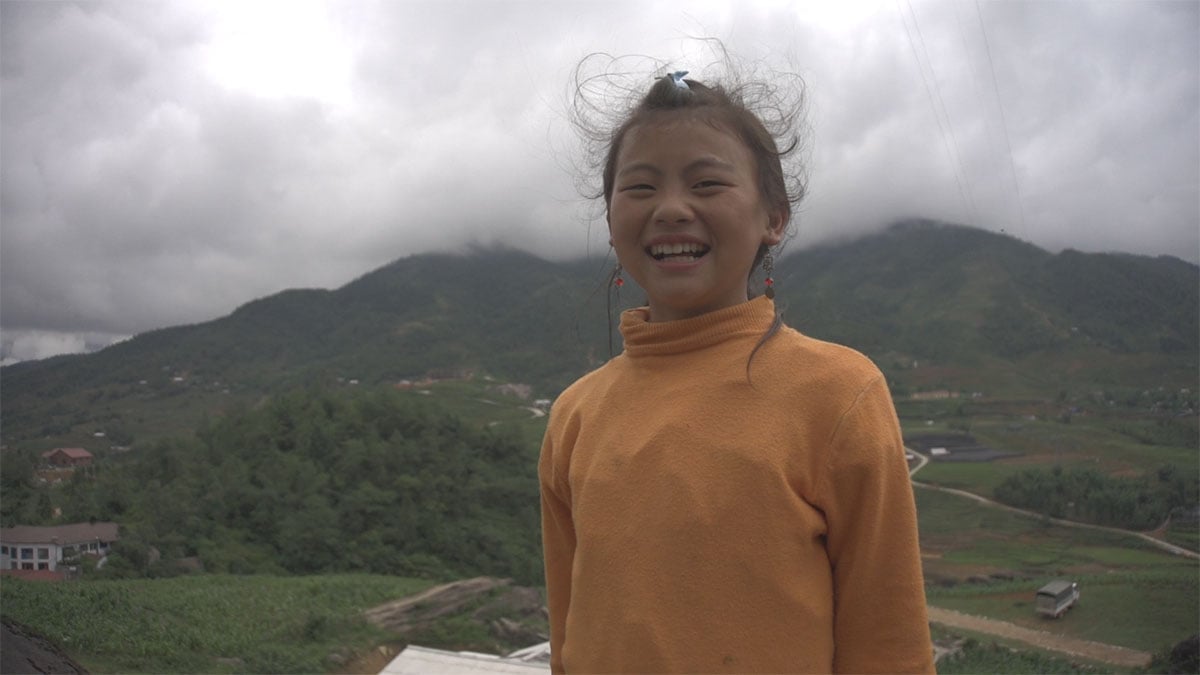 A 12-year-old girl is smiling widely at the camera. She is standing in the wind, wearing a yellow shirt. Behind her is a green, misty mountainside with a town below.