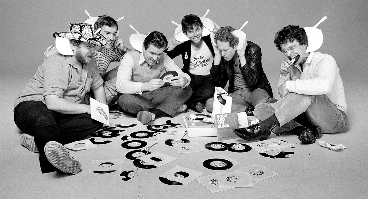 A black and white promotional band photo depicts six men sitting on the floor around a collection of vinyl records. They are playing with the paper album sleeves, and some have fitted them behind their ears to look like antennae. One album cover facing the camera reads “Doug and the Slugs.”