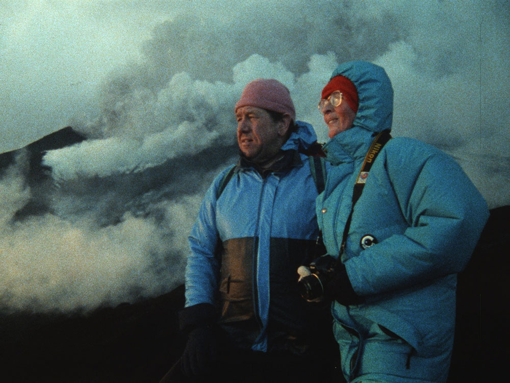 Two people stand on a mountainside as the dusk and clouds roll in. They are both wearing wearing blue winter jackets. Maurice, left, is in a pink tuque looking seriously into the horizon. Katia, right, is in a red tuque with her blue hood pulled up around it. She is wearing gold-rimmed glasses and smiling looking into the distance, holding a camera.