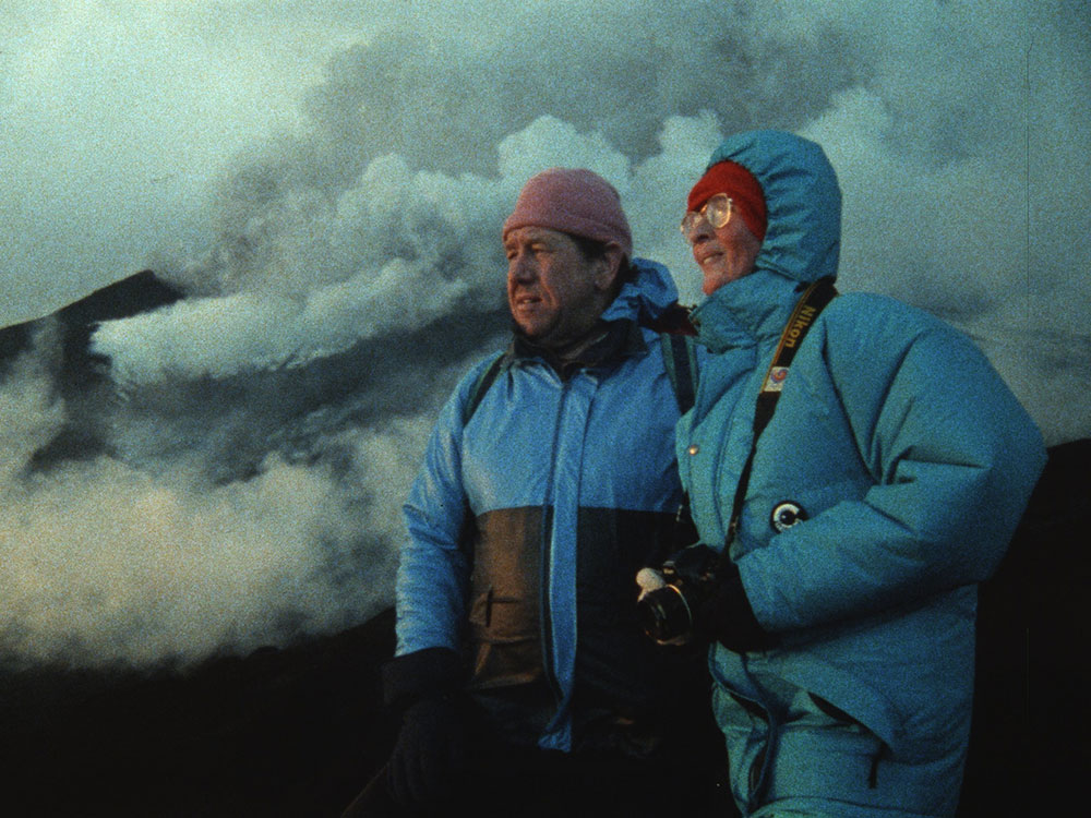 Two people in blue winter jackets are standing to the right of the screen, looking to the horizon on the left. Behind them, large white clouds of smoke from a nearby volcano billow around.