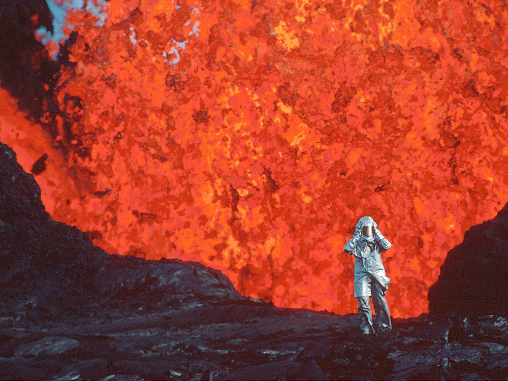 A person in a silver fire-proof suit holds their helmet in place with both hands as they walk towards the camera and away from a red-hot wall of magma from a volcano behind them.