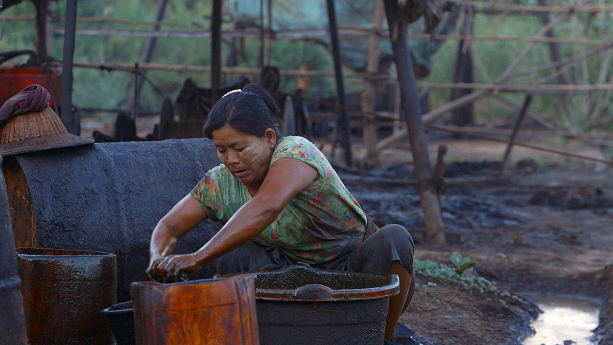 A woman crouches over a bucket outdoors, wringing out a cloth. The light is fading from the sky. Her hands and waist are stained with black oil.