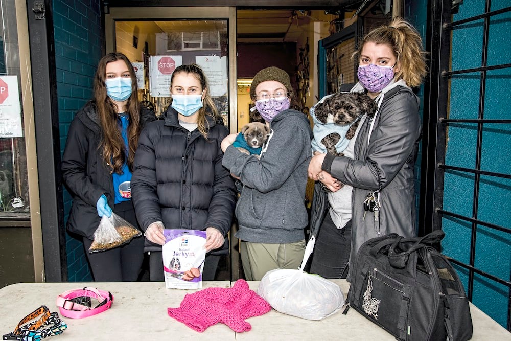 Charlie’s team, named above, stands behind a table holding leashes and sweaters. The team is wearing masks; two members are holding food and treats for animals, and two members are holding small dogs. 