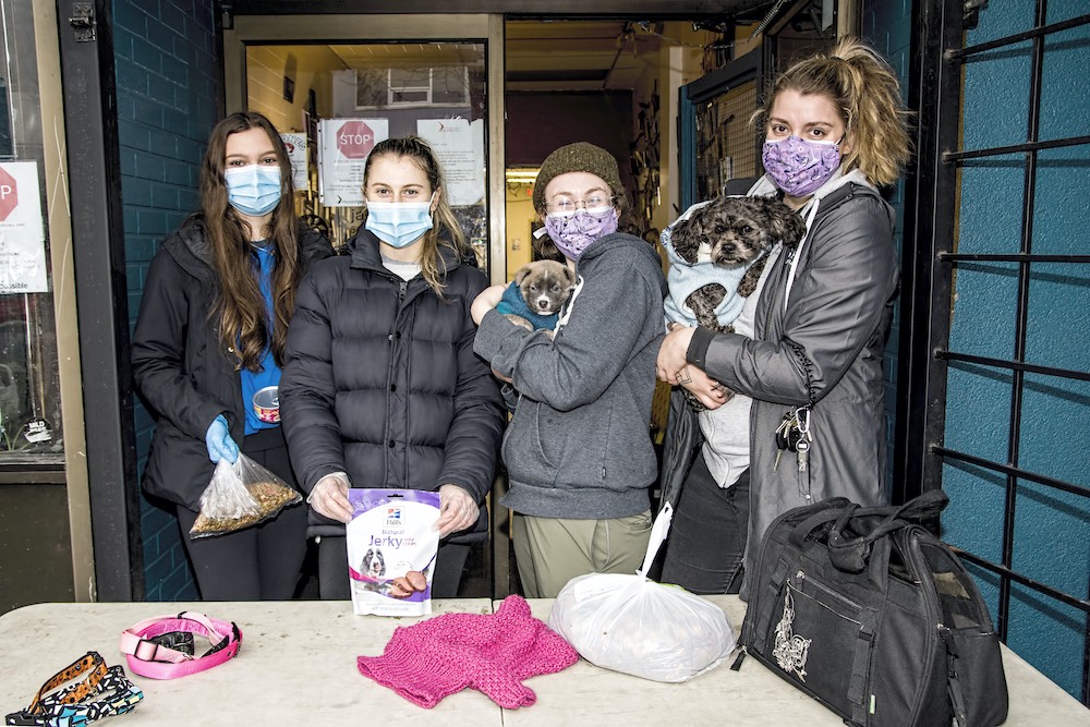 Charlie’s team, named above, stands behind a table holding leashes and sweaters. The team is wearing masks; two members are holding food and treats for animals, and two members are holding small dogs. 