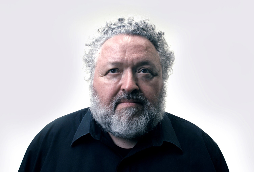 This is a shoulders-up portrait of designer Bruce Mau. He is a man with curly grey hair and a beard. He is looking up, away from the camera. He is wearing a black button-down shirt.