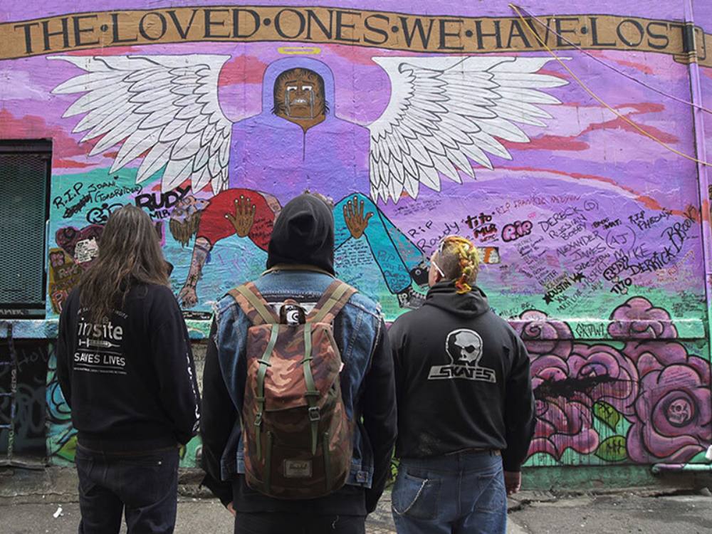 Three people stand next to each other with their backs turned to the camera. They are wearing black hoodies and denim. They are looking at a mural with a mauve background. Across the top, a hand- painted gold banner reads, “For the loved ones lost,” in memory of those who have died of toxic drug poisoning.