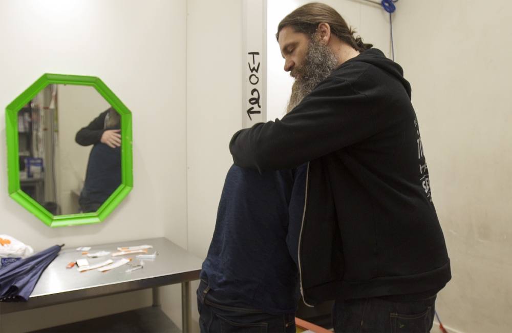 Ronnie Grigg is standing in a black hoodie. His long hair is tied back and he has a beard. He holds someone in his arms and is closing his eyes. Behind them, harm reduction supplies and an umbrella are scattered across a stainless steel table under a mirror with a green frame. 