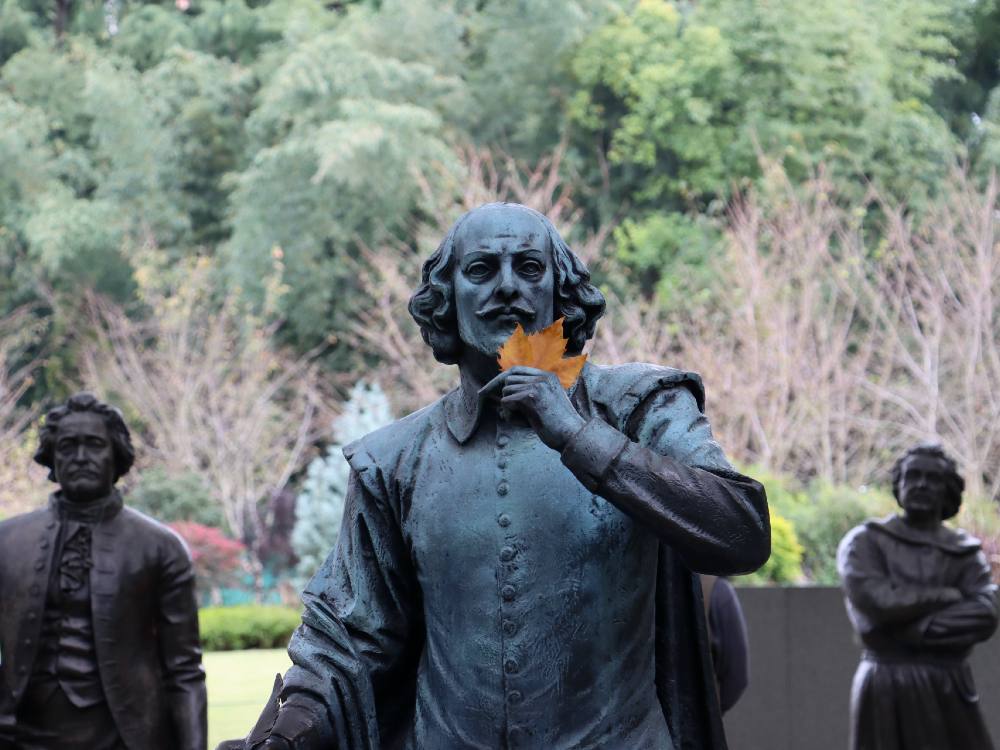 A statue of playwright and poet William Shakespeare holds a leaf to his face, flanked by other statues.