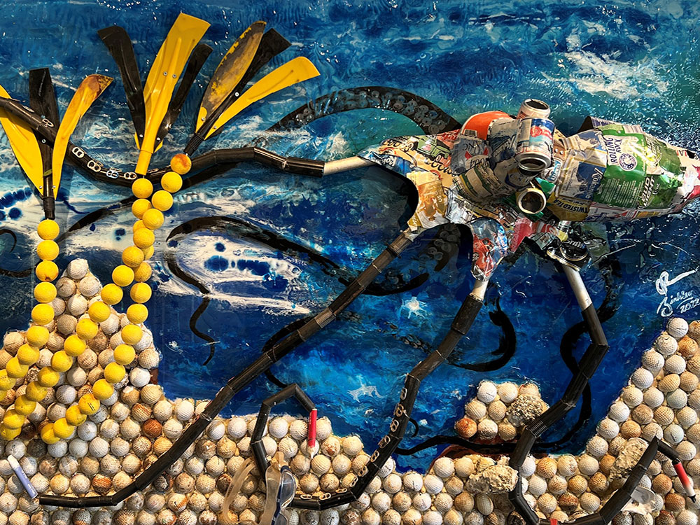 A whimsical representation of a squid with arms spreading in an undersea world. The squid is largely made of flattened beer cans and packaging, the eyes are made from ends of cans, and sand and sea plants are composed of golf balls. 