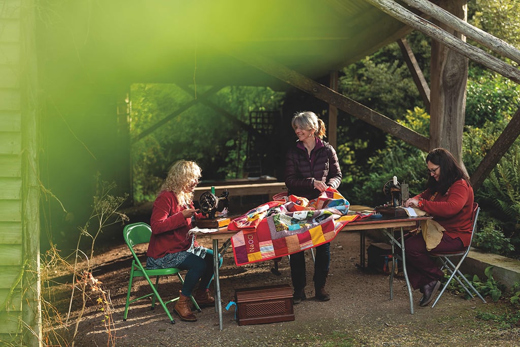 Three women work together at an outdoor table on a quilting project. Lush greenery frames the image.