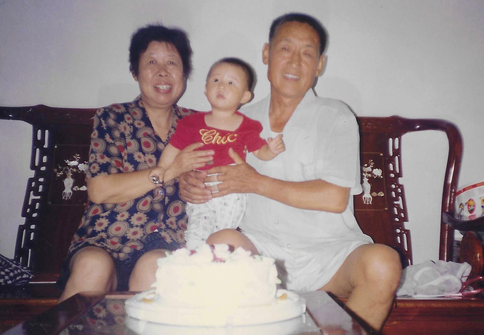 Author Jiaxuan (Josie) Wu is pictured here as a young toddler in the arms of her grandparents. Her grandmother is on the left in a dark blue patterned top. Her grandfather is on the right in a white short-sleeved button-down. The trio are seated on a wooden bench and a white cake is on the low table in front of them. 