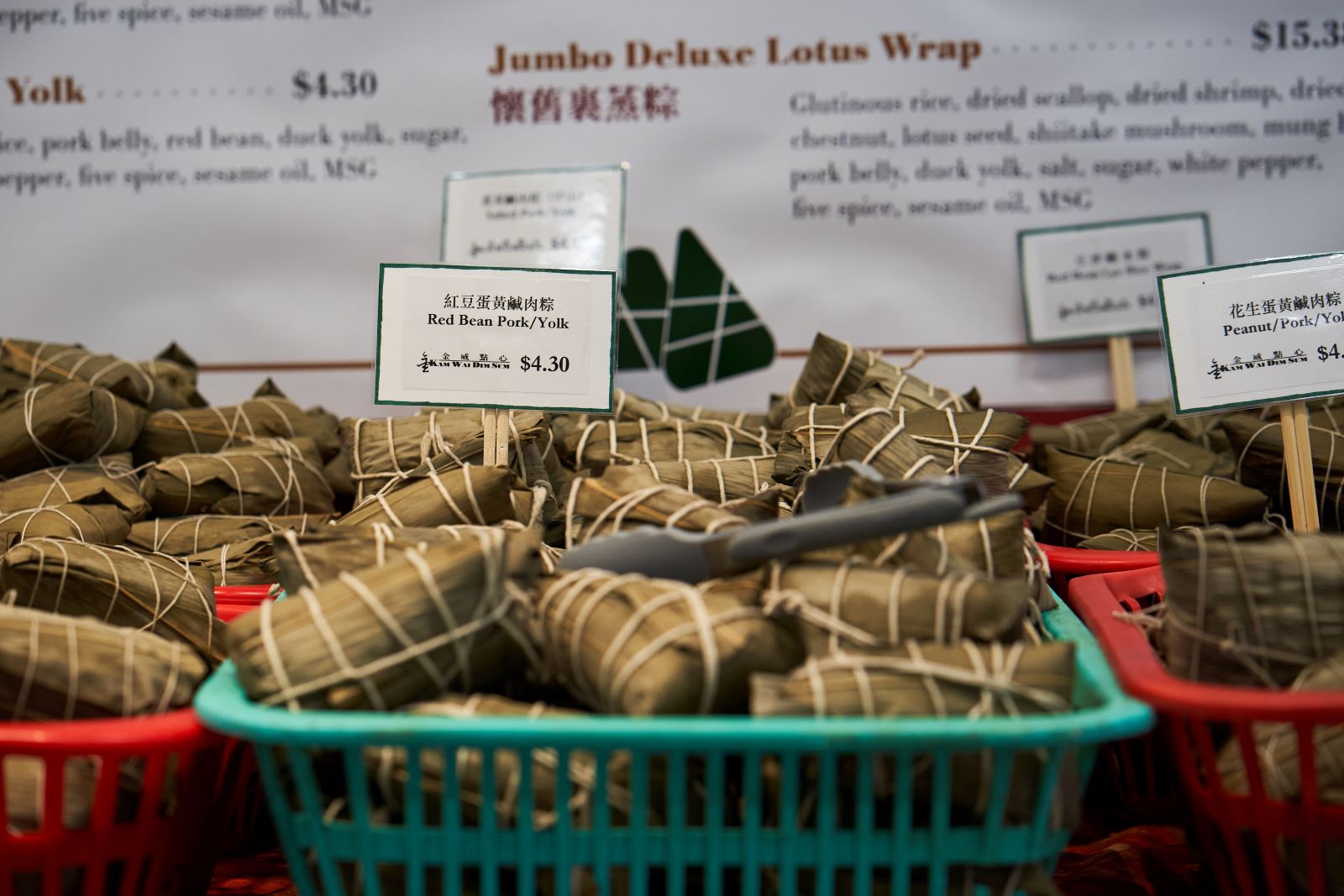 Red and green plastic bins overflow with zongzi at Kam Wai Dim Sum. The bins are arranged on a table with a large poster behind them describing each variety in detail.