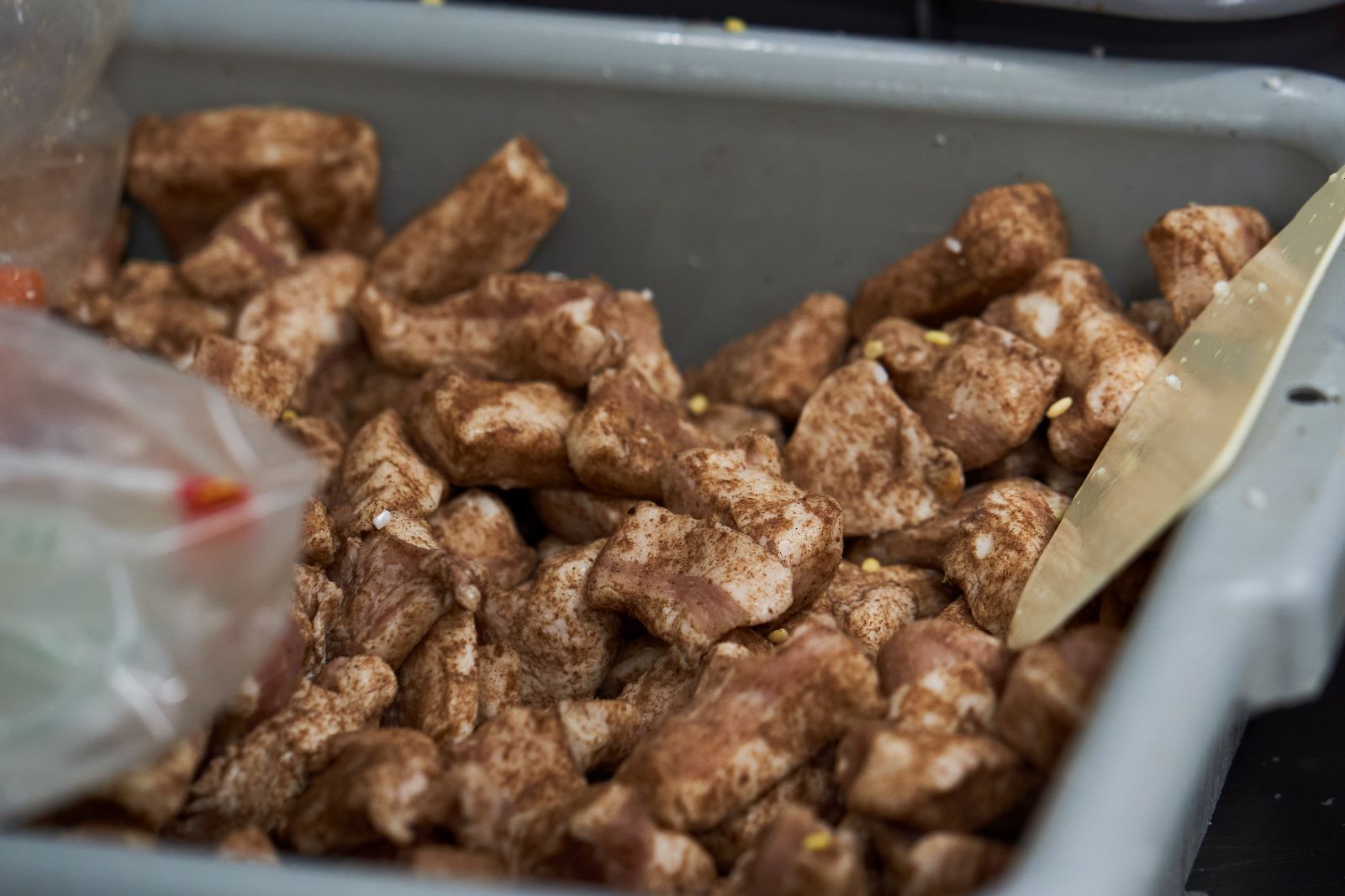 Pieces of pork belly are seasoned in dark brown spices in a grey plastic bussing tray.