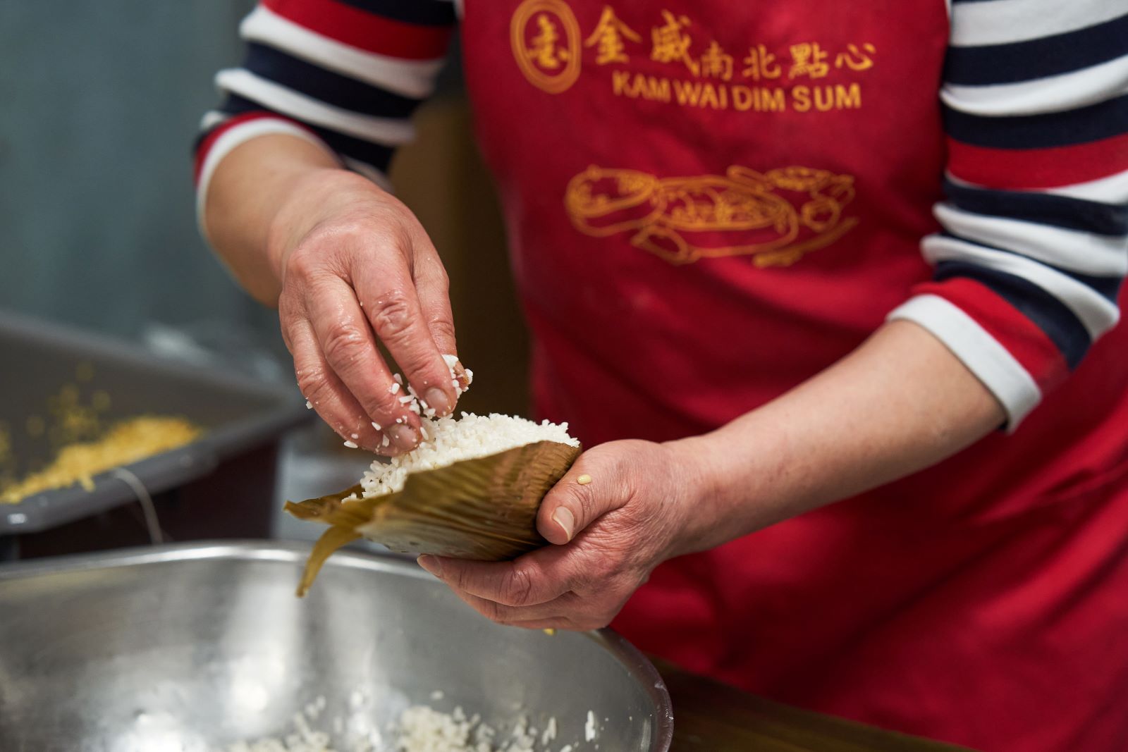 The hands of Xiao-Hong Wu stuff folded bamboo leaves with glutinous rice. Wu is wearing a red apron that reads ‘Kam Wai Dim Sum’ in yellow letters. Only Wu’s arms and apron are visible in this shot.