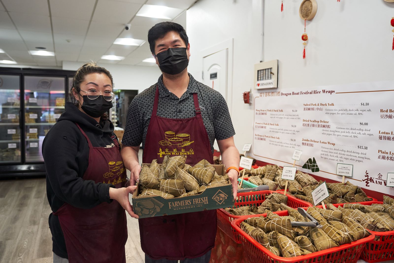 Susan (left) and William Liu (right) hold a box of zongzi at Kam Wai Dim Sum. They are wearing black medical masks and matching maroon apronx. They are looking at the camera and smiling.