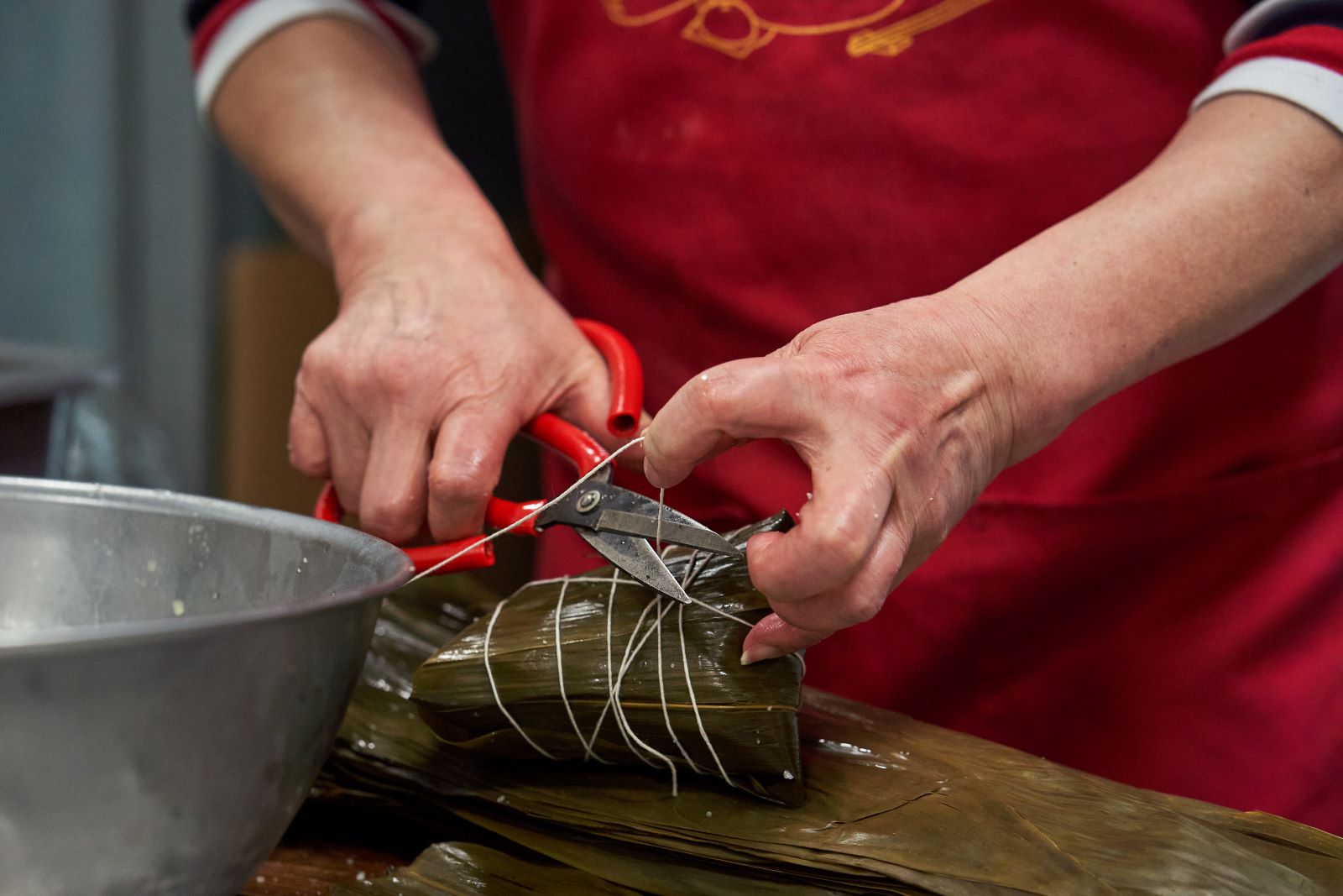 The hands of Xiao-Hong Wu trim butcher’s twine around a zongzi with a pair of red kitchen scissors. Wu is wearing a red apron and is cutting the zongzi at a counter on which a metal mixing bowl sits in the foreground. Only Wu’s arms and apron are visible in this shot.