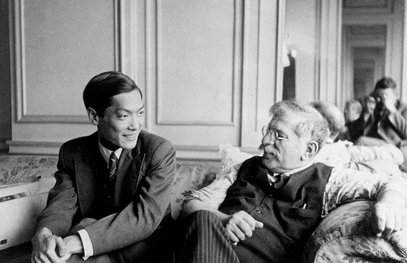 A black and white photo of a young Li Shiu Tong, left, and an older Magnus Hirschfeld, right, sitting on a floral soft. There is a mirror behind the sofa and the photographer is visible in the mirror.