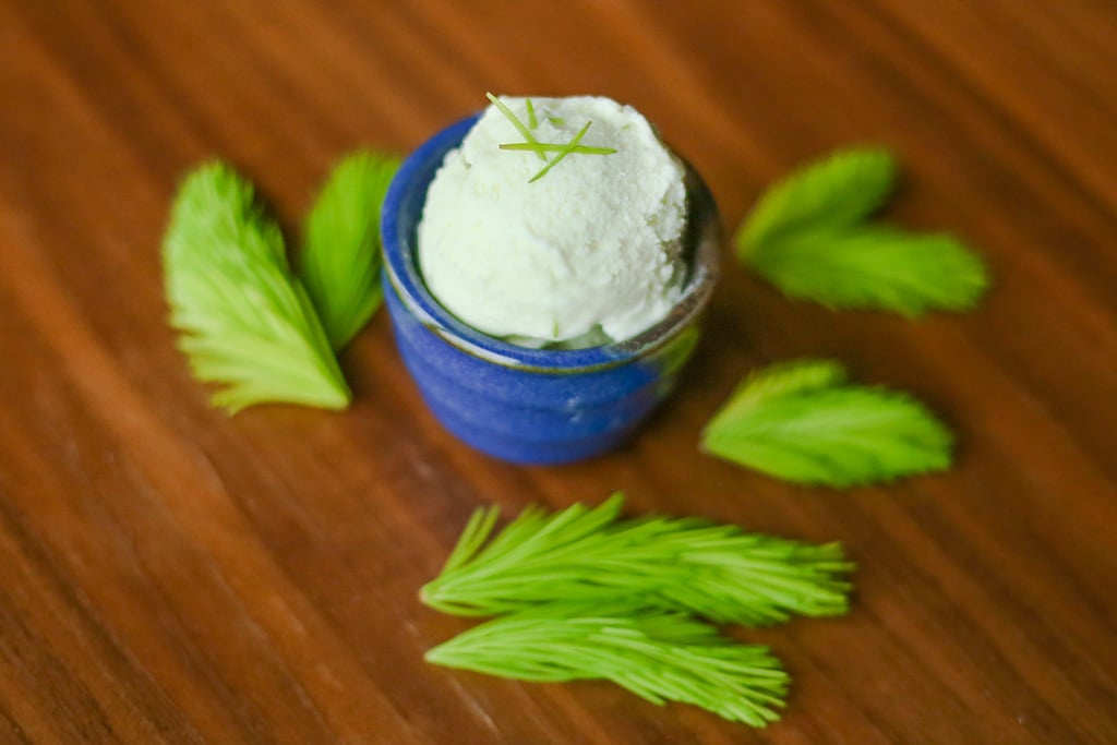 A small blue cup with a scoop of green-tinted ice cream on a wooden table, with spruce tips scattered about.