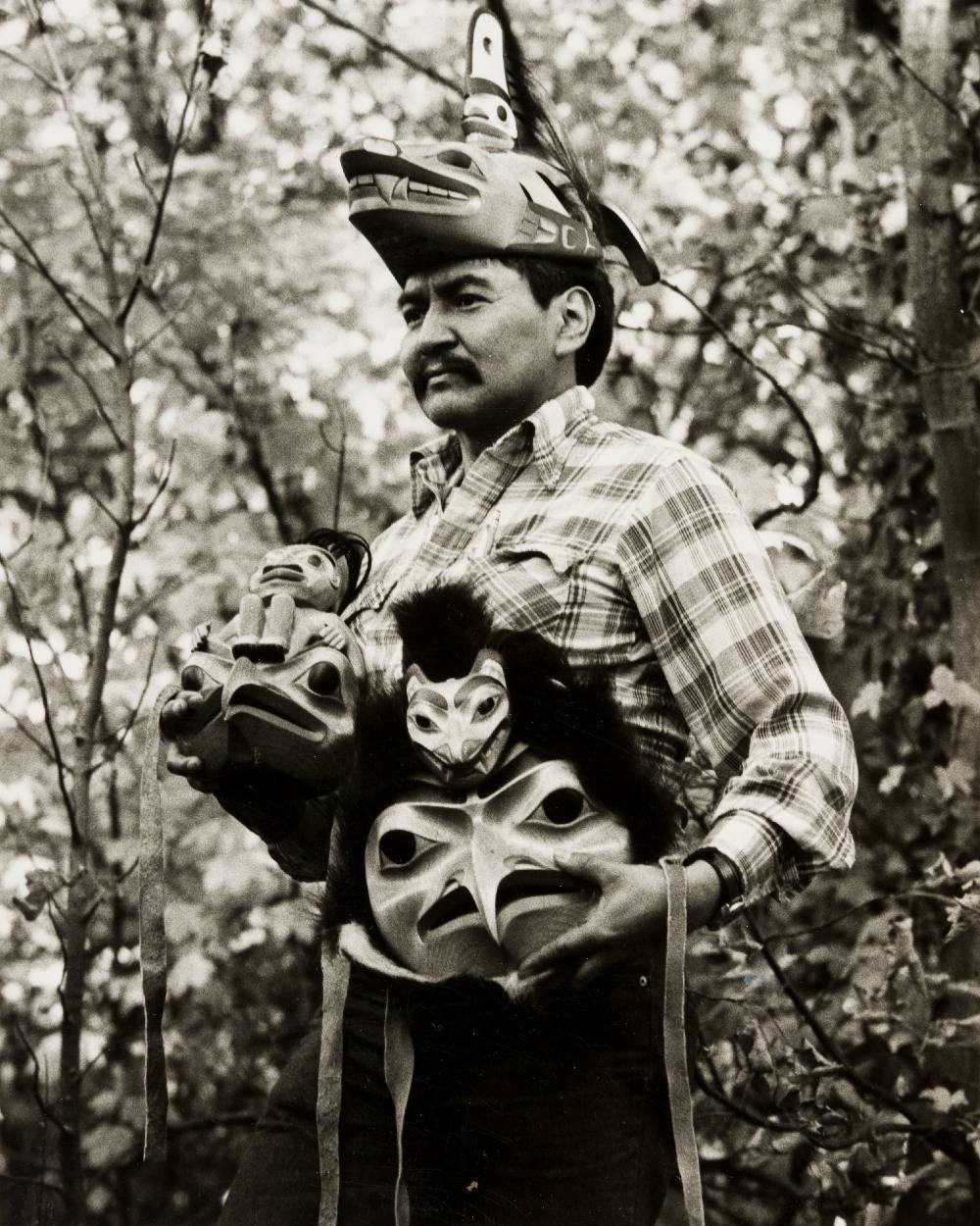 A black and white photo of Dempsey Bob depicts the artist wearing and holding three of his works from 1987. He is standing in a plaid shirt. He is wearing <em>Killer Whale Headdress</em> and holding <em>Eagle Human Mask</em> and <em>Eagle Bear Mask</em>. He is looking towards the left side of the frame with trees behind him. 