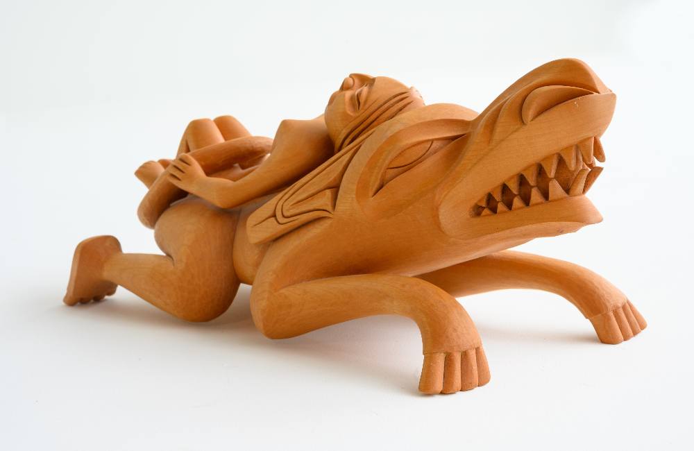An alder wood sculpture of a woman lying face-up on the back of a wolf. The wolf is looking towards the right of the frame. Its snout and sharp teeth are prominent.