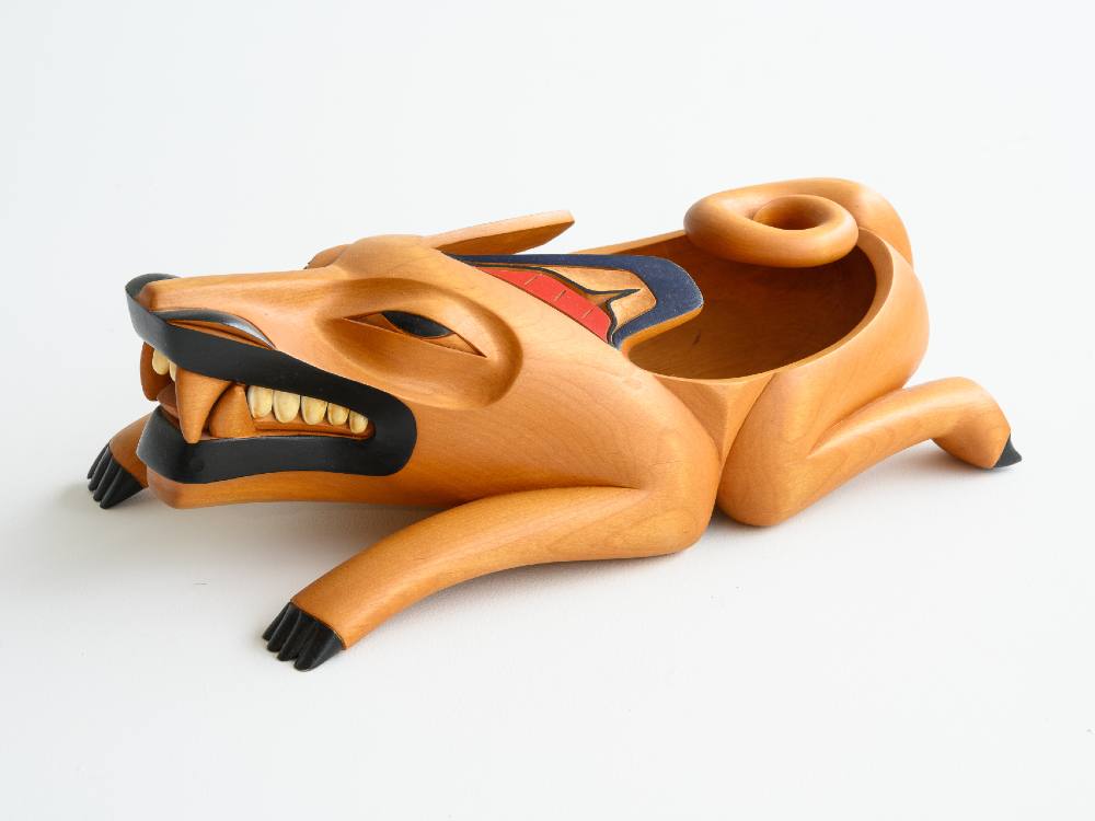 A wooden sculpture of a wolf uses golden alder wood and black and red acrylic paint. The wolf’s head and teeth are towards the left of the frame. Its sharp teeth are visible and emphasized by black paint on its lips.