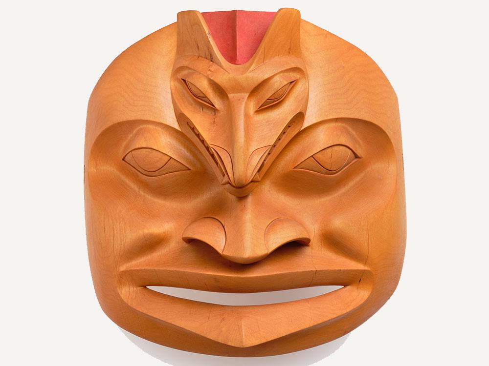 A wooden mask in a rich golden tone is made of alder wood with a touch of red acrylic paint. The mask depicts a wolf’s face on the forehead of a human face.
