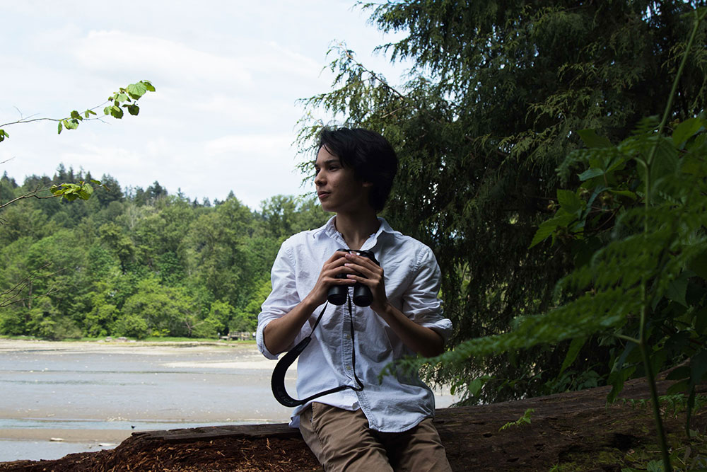 A teenager wearing a blue collared shirt sits on a log, looking to his right, holding a pair of binoculars. 