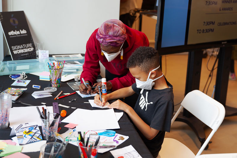 A child sits at a table full of drawing materials, scissors and glue. They are using a pen and writing or sketching on some paper. An adult stands beside them, working on their own project.