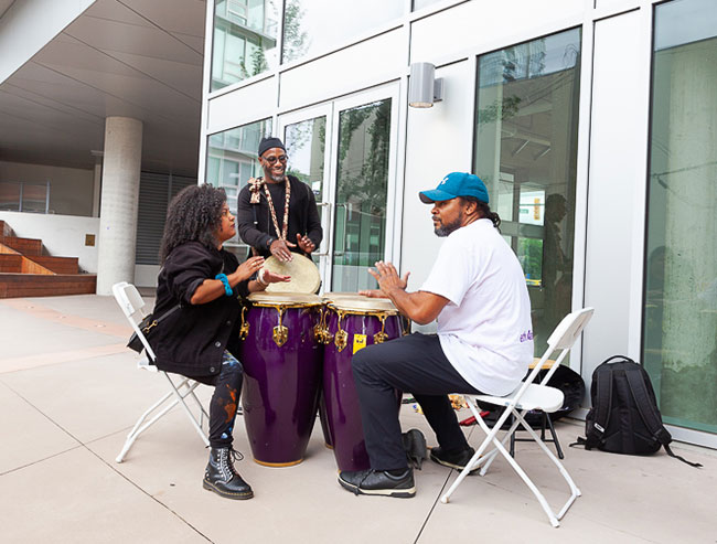 Three people play drums. They are outdoors.