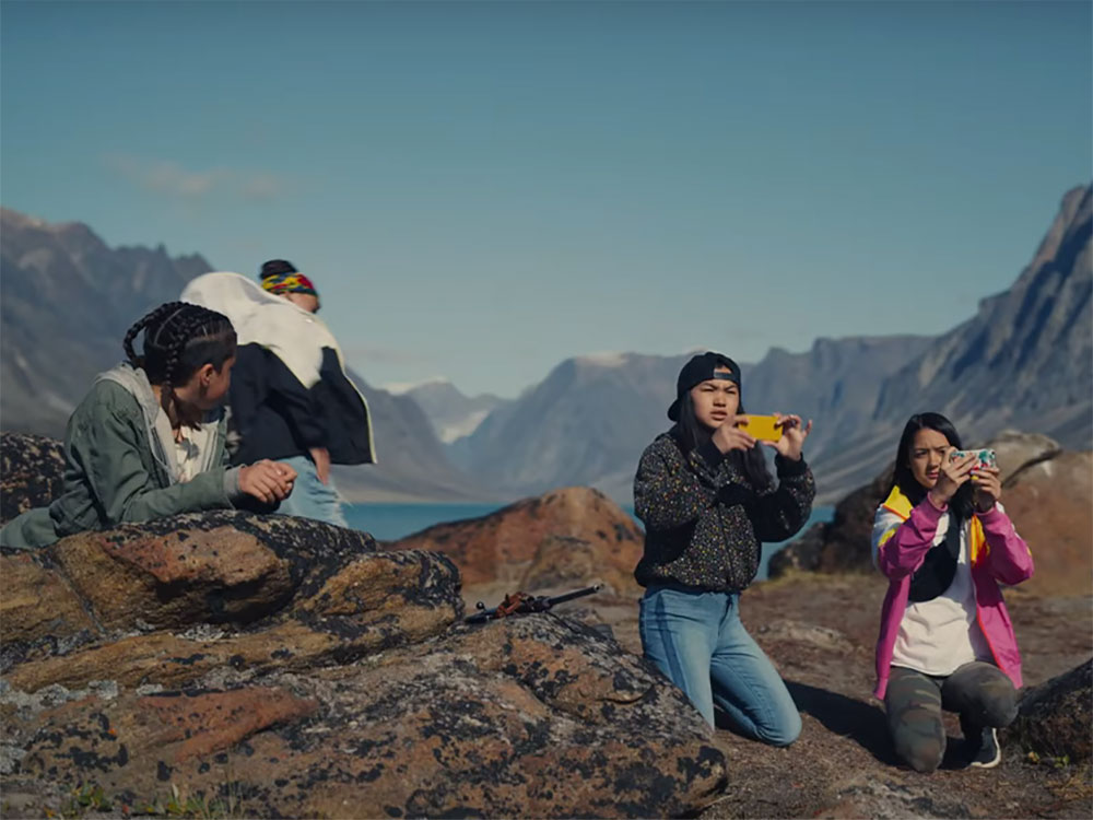 A group of four teens hold up their phones to look at something out of frame in the horizon. Behind them is a spectacular mountain range and lake.