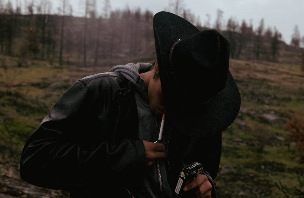 A young man, Elijah Swan-Hall, lifts a lighter to the cigarette in his mouth, his face half-hidden by the brim of a black cowboy hat. 
