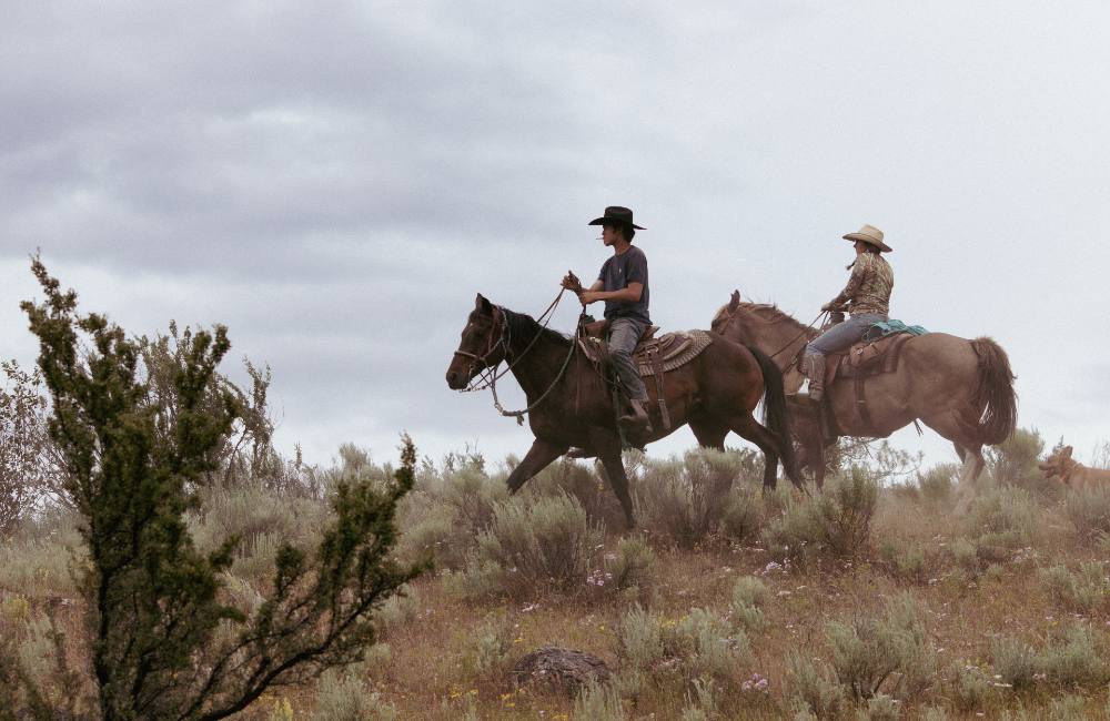 Jessi Wyatt and Elijah Swan-Hall, mounted, walk across the ridge of a hill covered in sagebrush. A cigarette hangs from Swan-Hall’s mouth as he lifts the reins.