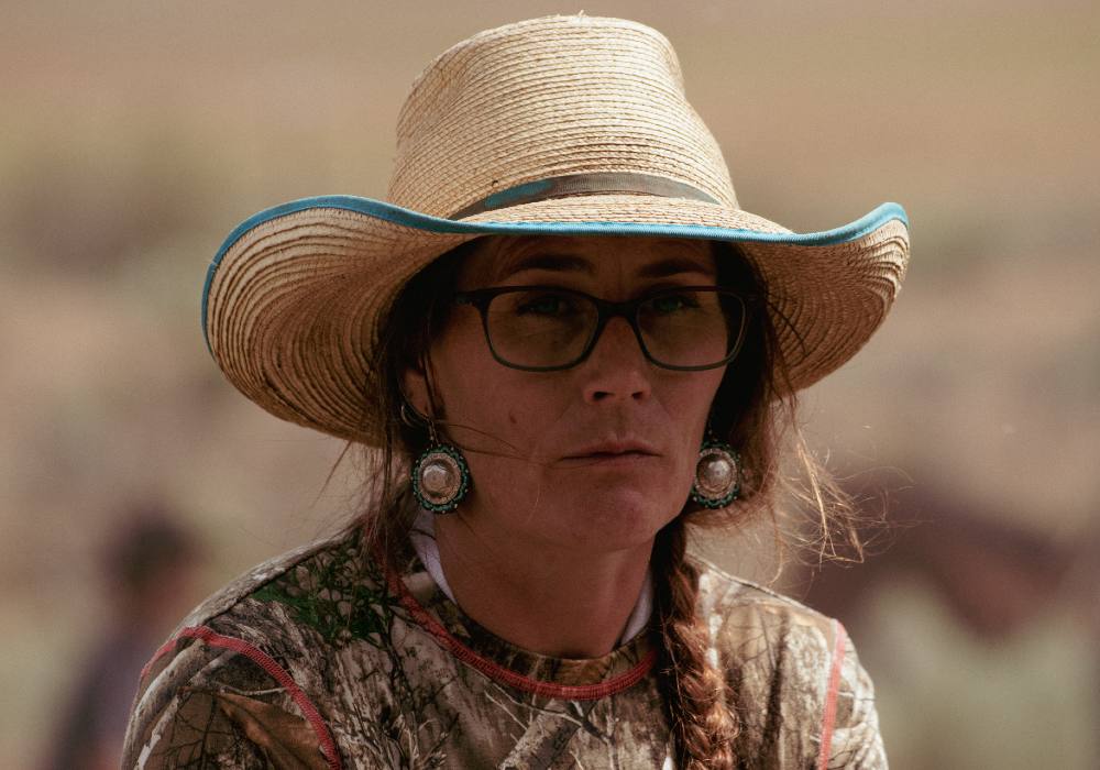 A woman, Jessi Wyatt, looks past the camera. A worn straw hat shadows her face from the sun. She is wearing turquoise and silver medallion earrings. Wisps of hair have escaped her braid and frame her face. 