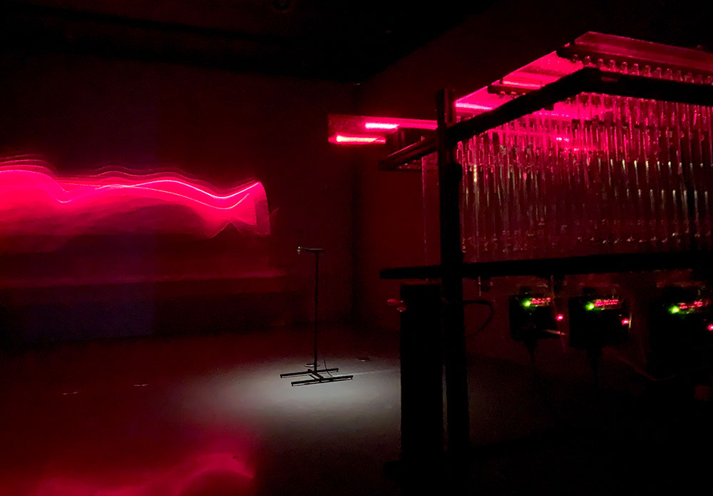 Bright red projections cut through darkness. To the right of the frame, an electronic panel is visible.
The exhibition’s introduction places the individual pieces in the show into this human context: “Ancient cosmology was rooted in our meticulous observation of natural patterns. Hundreds of thousands of years in the making, the most sensitive, sophisticated, and flexible instrument of observation may still be us; our frail bodies and creative minds.” 