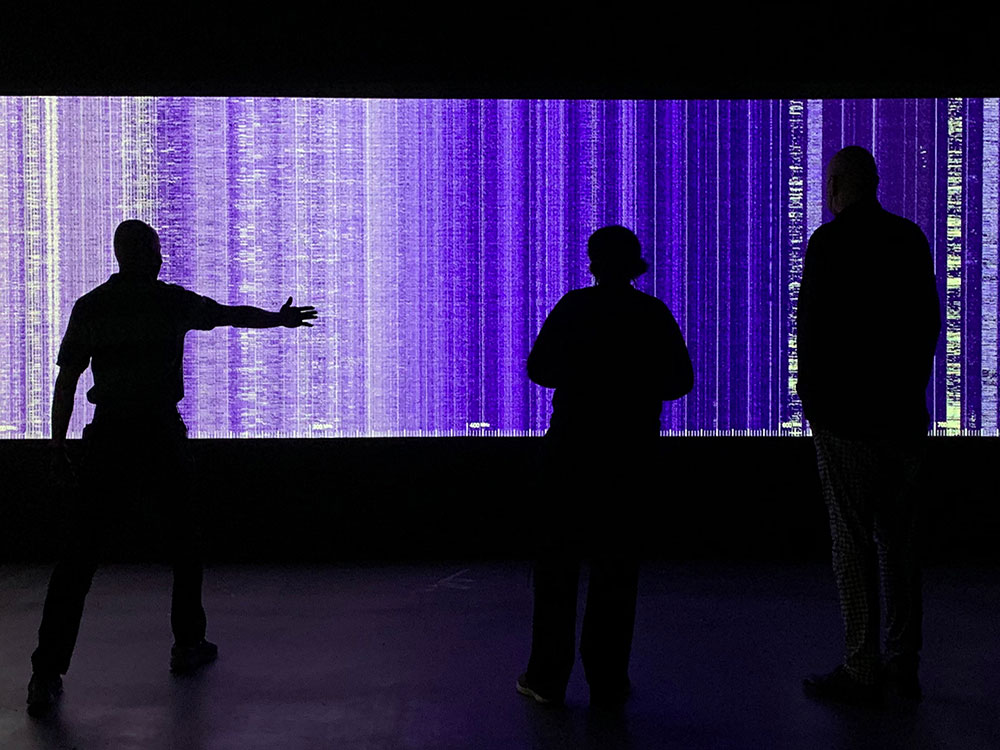 The black silhouettes of three people can be seen in front of a prismatic indigo digital screen depicting the radio spectrum.