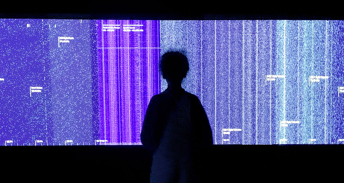 A person stands in front of a prismatic indigo screen depicting the radio spectrum.