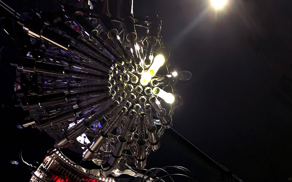 A piece of industrial design features silver Gelger Müller tubes, glass, aluminum and micro controllers. The piece is large, silver and hangs from the ceiling of the New Media Gallery.