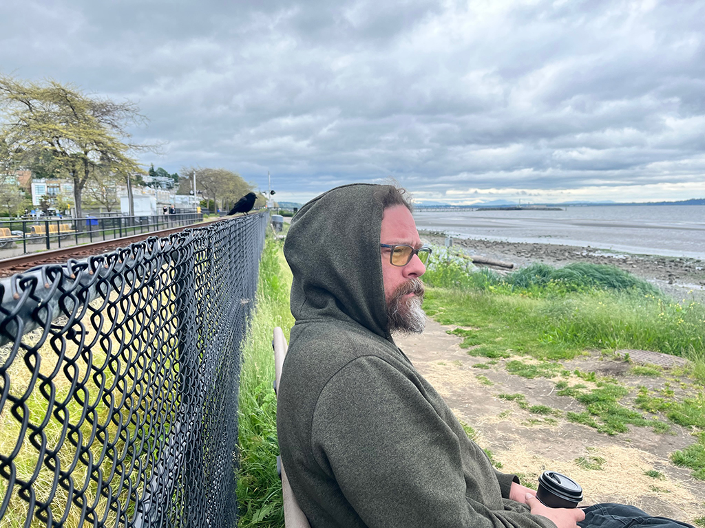 Joseph Kakwinokanasum is wearing a grey hoodie and and glasses. He is holding a black coffee cup and sitting on a bench in front of a black chain-link fence to the left. To the right is a beach and the ocean.