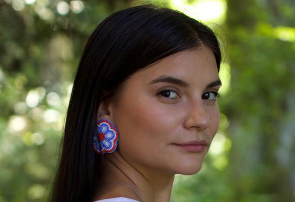 Selina Boan turns her head to look at the camera. She is wearing a colourful beaded earring in the shape of a flower. She has long dark hair. There is greenery in the background. 