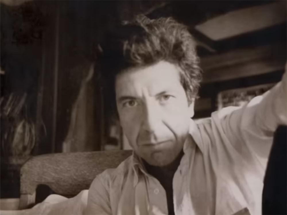 A sepia-toned photo of Leonard Cohen shows the singer seated on a sofa and looking directly at the camera. His expression is serious.