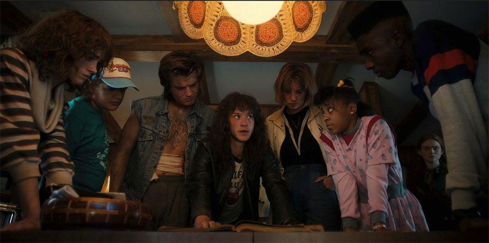 Seven teens circle a table. The teen in the middle is reading from a book on a table. Overhead, a crocheted light is visible.